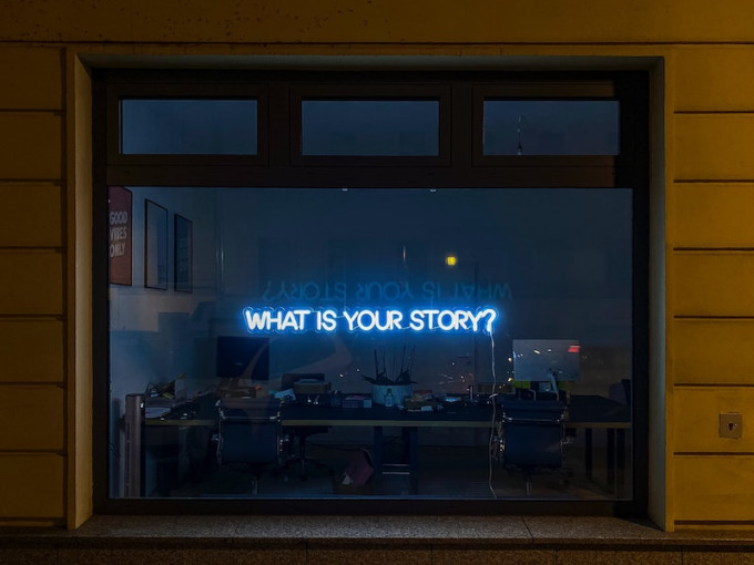 Neon sign in office window with the words what is your story written on it