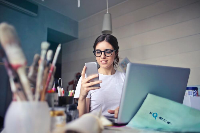 Young woman sitting at desk with laptop open looking at her smartphone