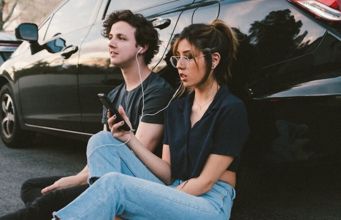 Guy and girl sitting on the ground outside their car listening to a podcast