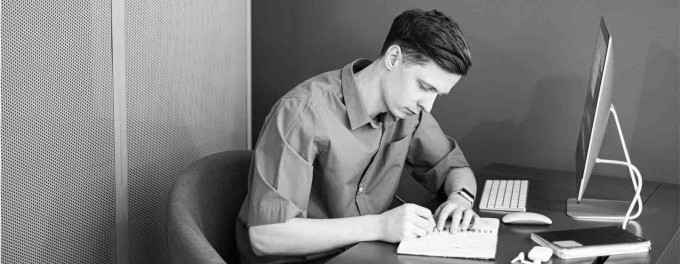 Young guy sitting at desk with a computer, notebook and pen, writing a podcast description