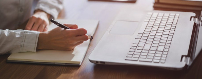 Closeup of person writing in a notebook with a laptop open in front of them