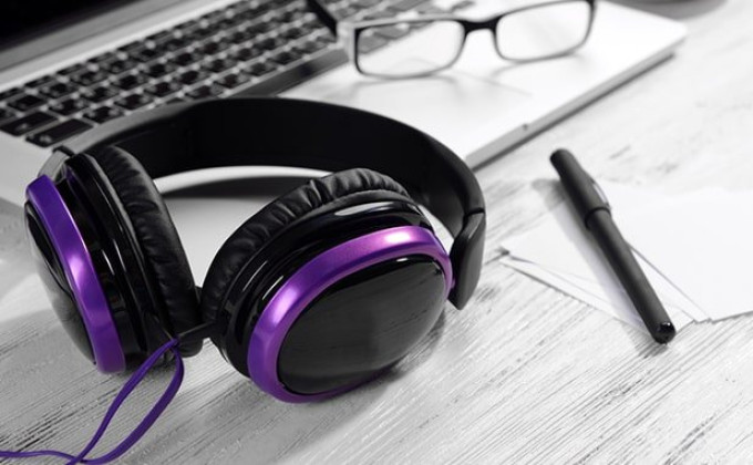 Image of black headphones with purple trim lying on a desk with a keyboard and glasses