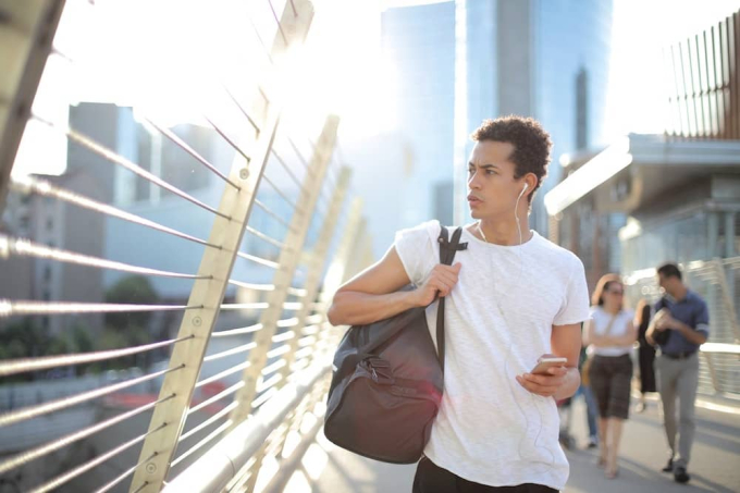 Young guy walking with kit bag, listening to a podcast on his smartphone